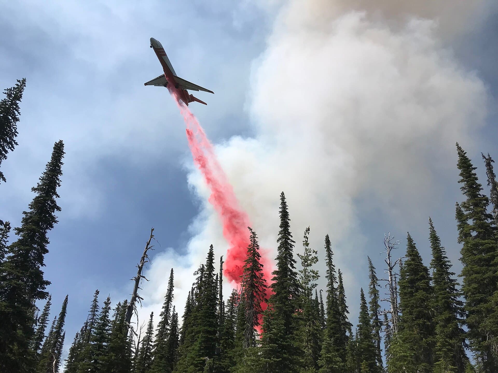 Airtanker releasing a long stream of bright red retardant above a smoke-filled forest